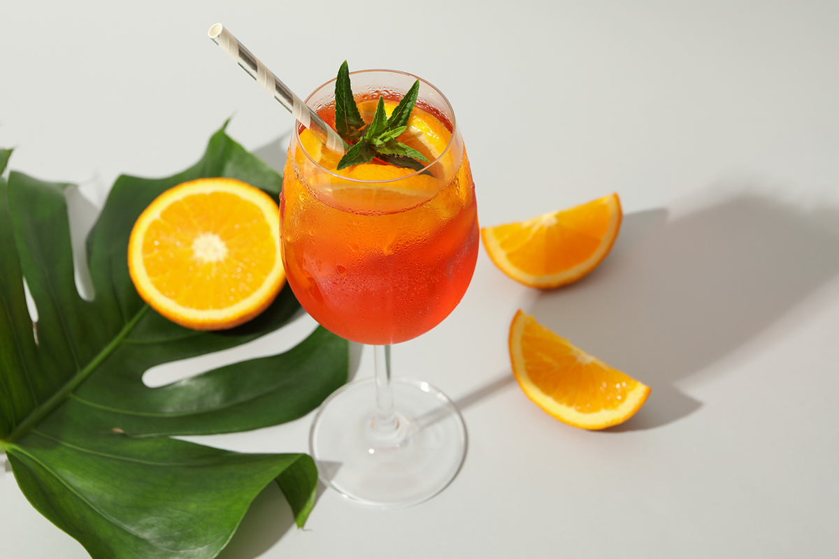 Komposition mit Aperol Spritz Cocktail auf weissem Hintergrund mit Monstera und Orange.