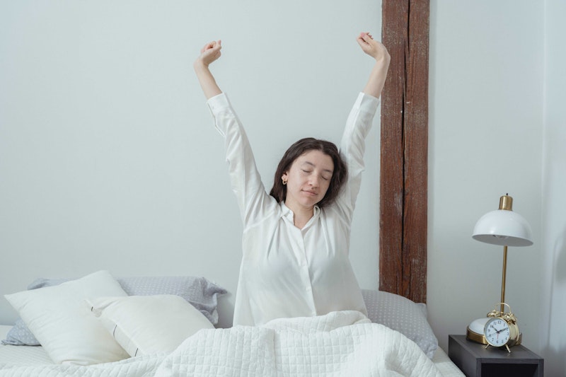 Eine Frau in weissem Schlafanzug sitzt aufrecht im Bett und streckt ihre Arme nach oben. 