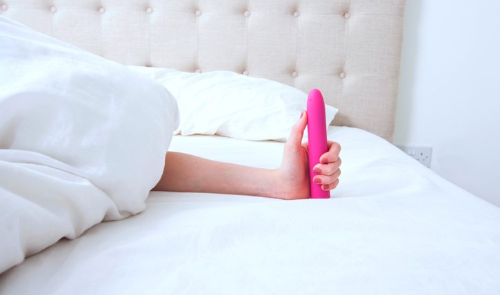 Eine Frau im Bett hält einen Vibrator in die Luft.