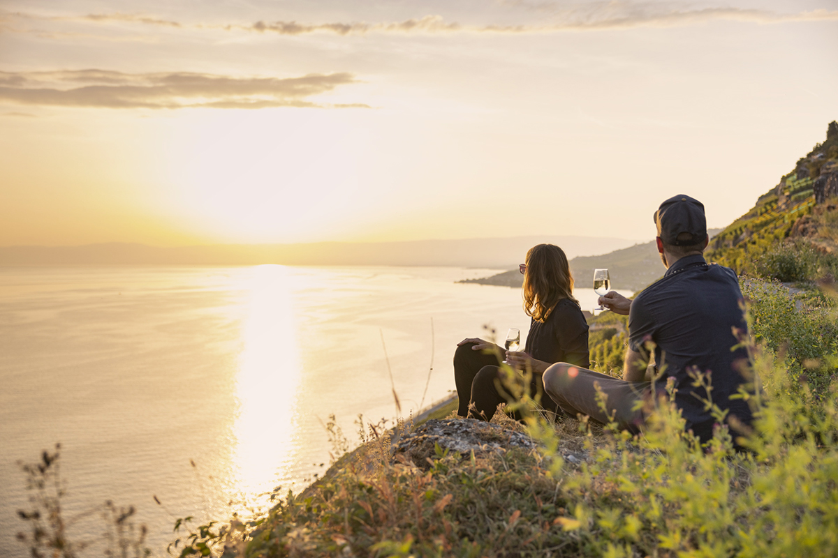 Ein Paar sitzt in den Lavaux-Rebbergen und geniesst mit einem Glas Wein den Sonnenuntergang mit Blick auf dem Genfersee.